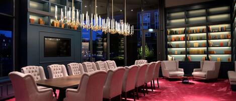 A modern dining area with a long table and upholstered chairs, a chandelier, and bookshelves with books.