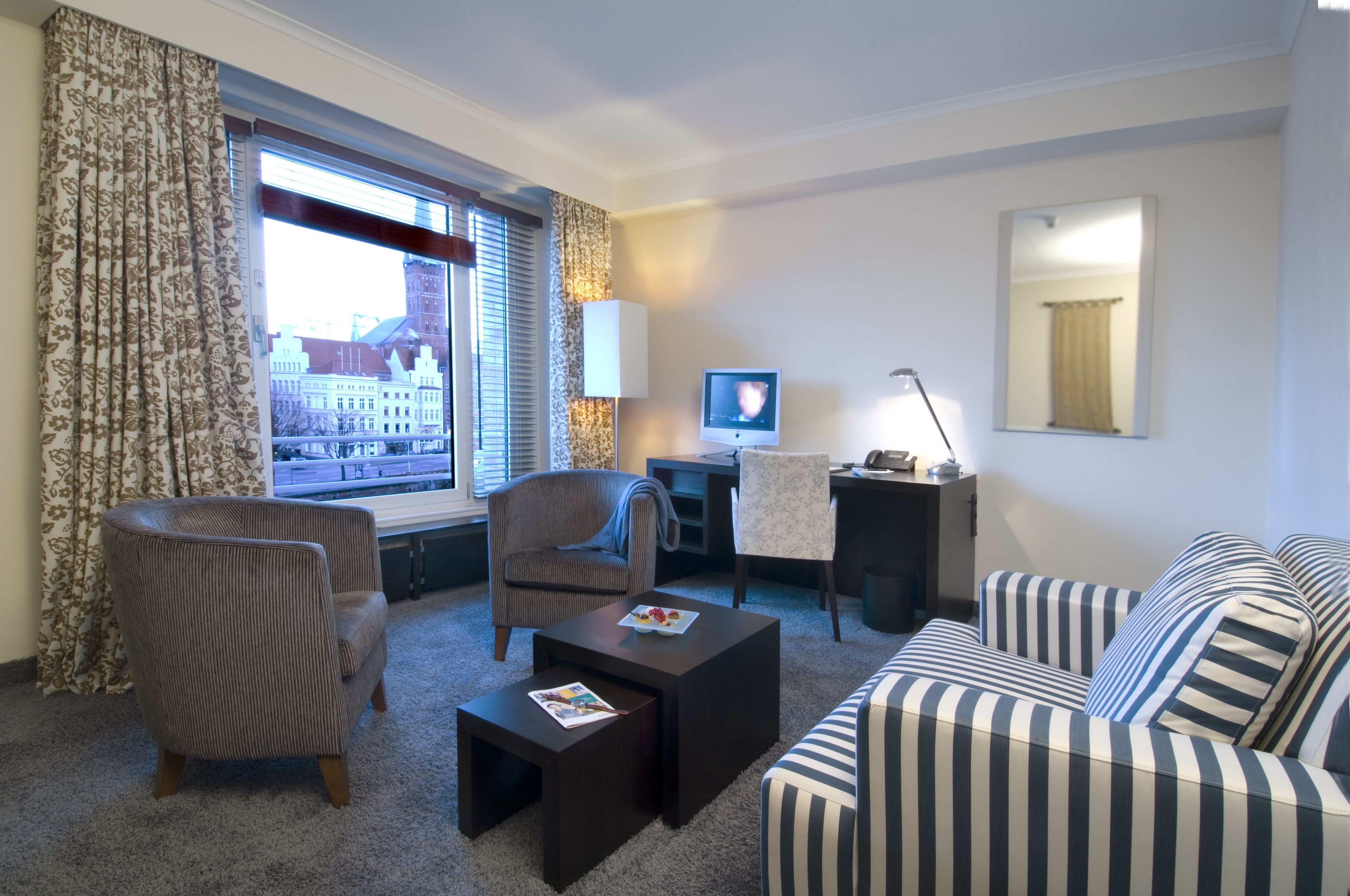 A hotel room with a view of a cityscape, a striped sofa, a striped armchair, a small table, and a desk with a lamp.