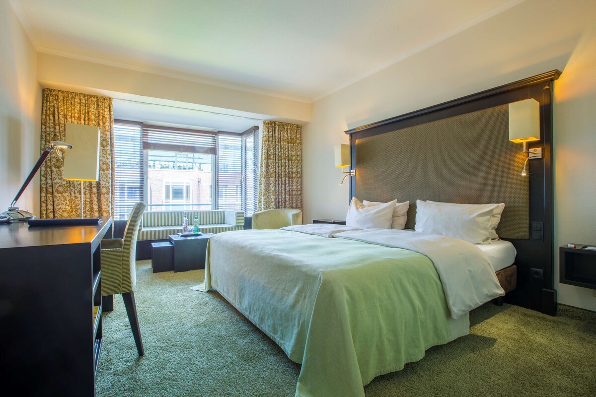 Ein Hotelzimmer mit Bett, Schreibtisch, Stuhl und Blick auf die Stadt durch große Fenster.