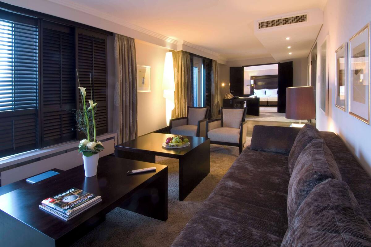 A modern hotel room with a dark brown sofa, a coffee table with a fruit plate, a white vase with flowers, and a desk with a remote control and magazines.