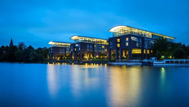 A modern hotel with a glass facade, located by a body of water. - Radisson Blu Senator Hotel (Lübeck)