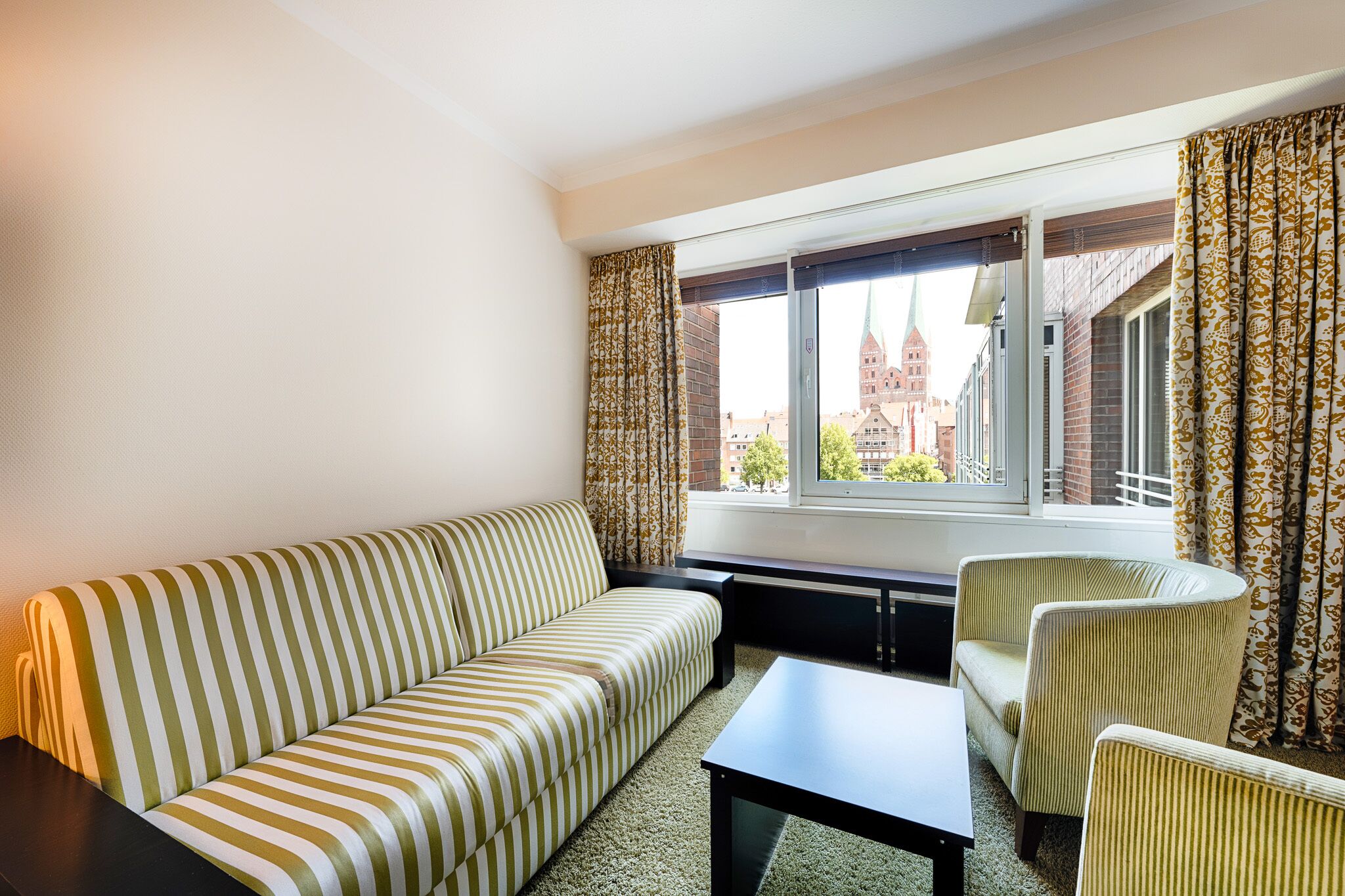 Ein Zimmer mit einem gestreiften Sofa, einem kleinen Tisch und zwei Sesseln. Ein Fenster mit gemusterten Vorhängen bietet Blick auf ein Gebäude mit einem Kirchturm.