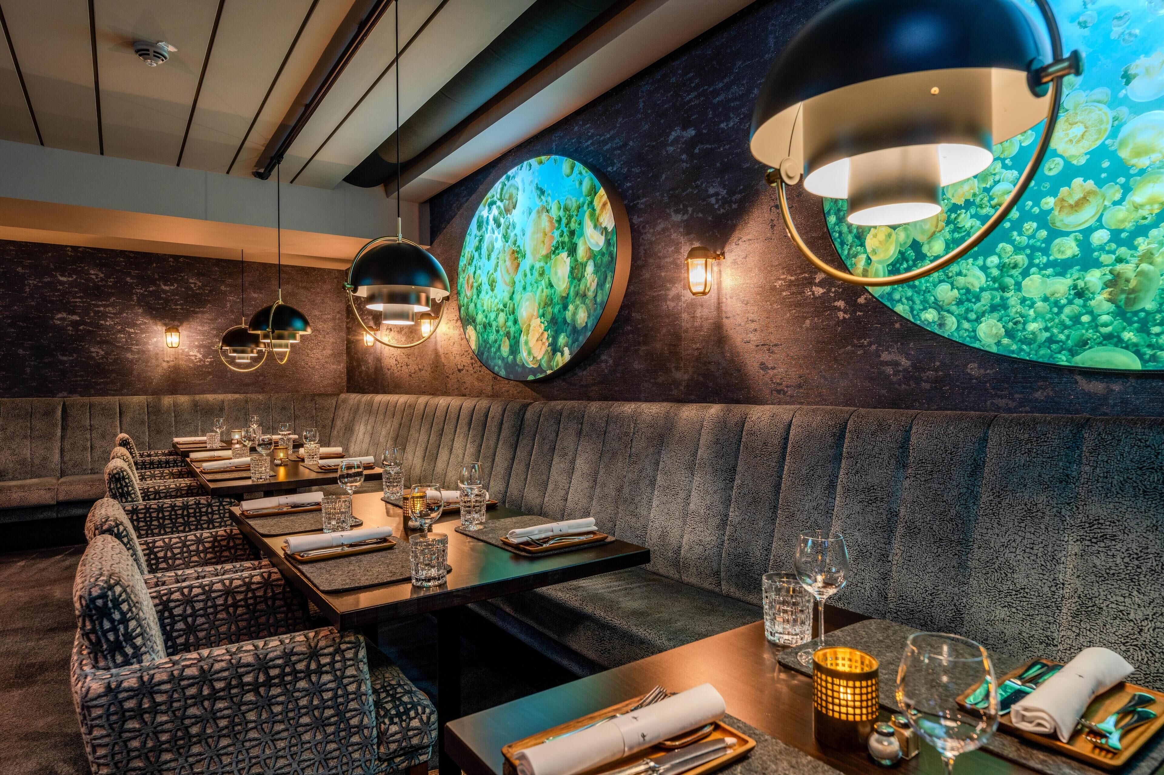 A restaurant with a modern interior, featuring a long dining table with multiple chairs, a cushioned bench, and a wall with circular decorative elements.