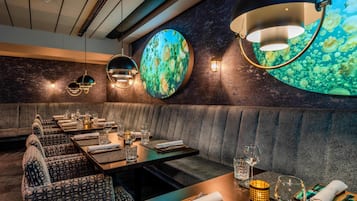 Un restaurant au décor moderne, avec une longue table à manger entourée de nombreuses chaises, un banc rembourré et un mur orné d’éléments décoratifs circulaires.