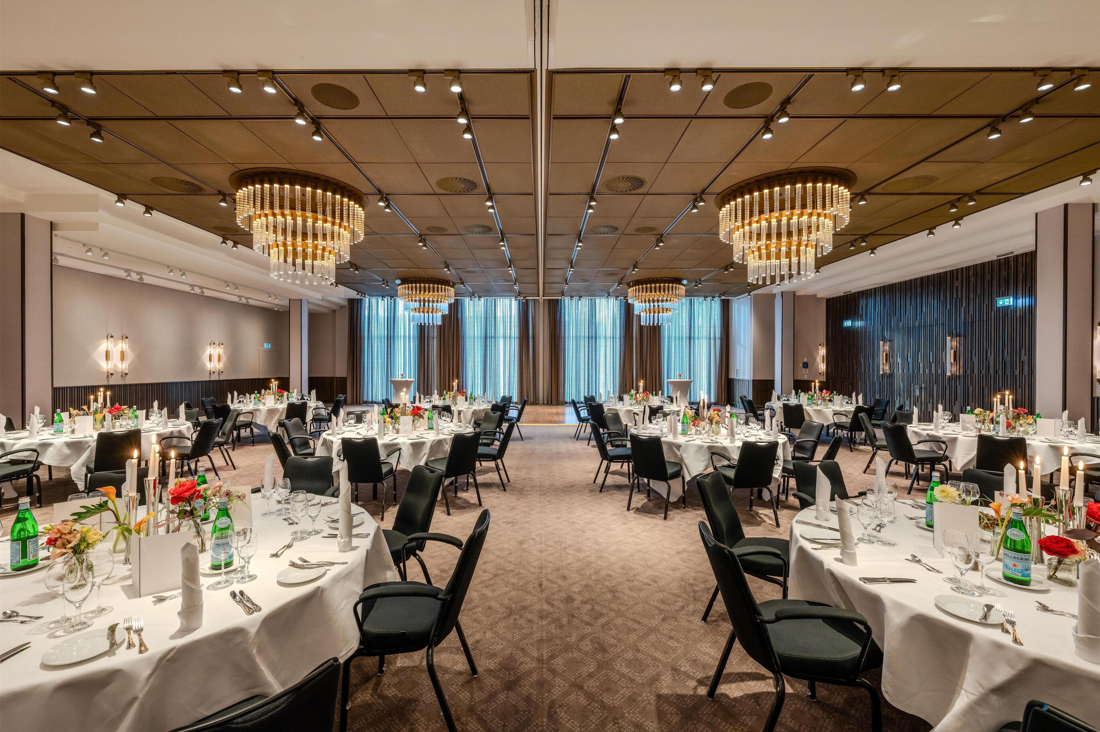 Ein geräumiger Festsaal mit mehreren runden Tischen, gedeckt für ein Essen, mit weißen Tischdecken, schwarzen Stühlen und stilvollen Tischdekorationen.