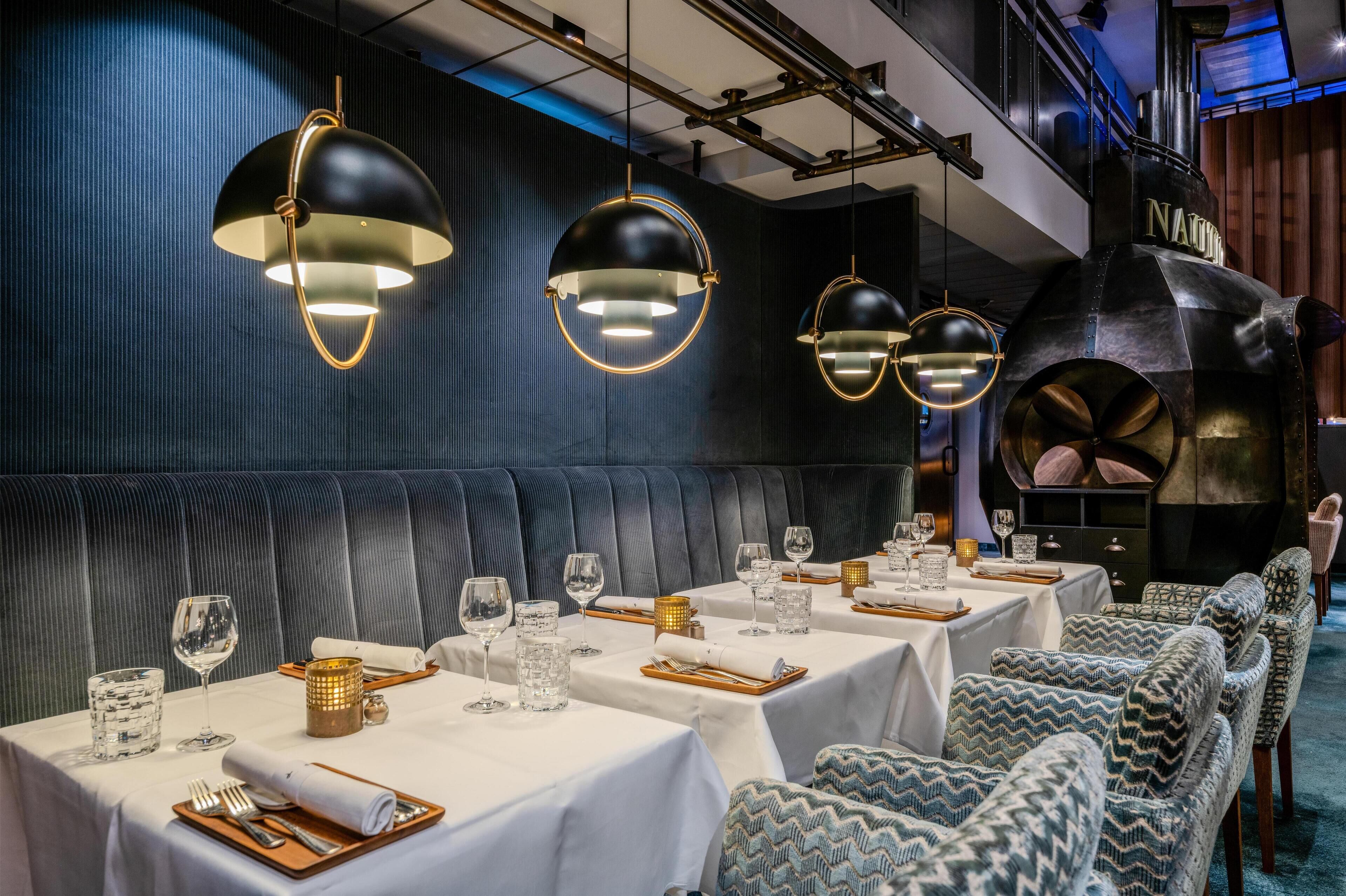 Un restaurante con varias mesas dispuestas para comer, iluminación colgante negra y un horno de leña al fondo.