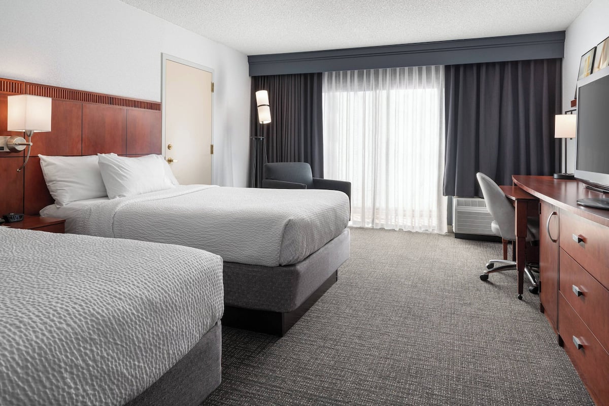 Habitación de hotel con dos camas, un escritorio, una silla, un televisor y un ventanal con cortinas.