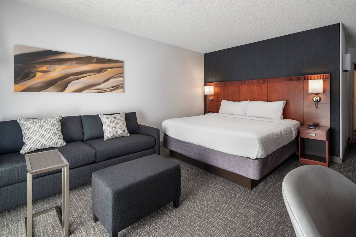 Una habitación de hotel con una cama, un sofá, un reposapiés, una mesita y una mesita de noche con una lámpara.