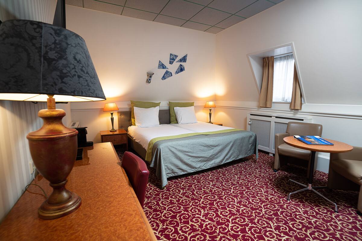 A hotel room with a bed, two bedside lamps, a desk with a lamp, a chair, and a small table with a book.