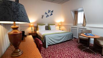 A hotel room with a bed, two bedside lamps, a desk with a lamp, a chair, and a small table with a book.