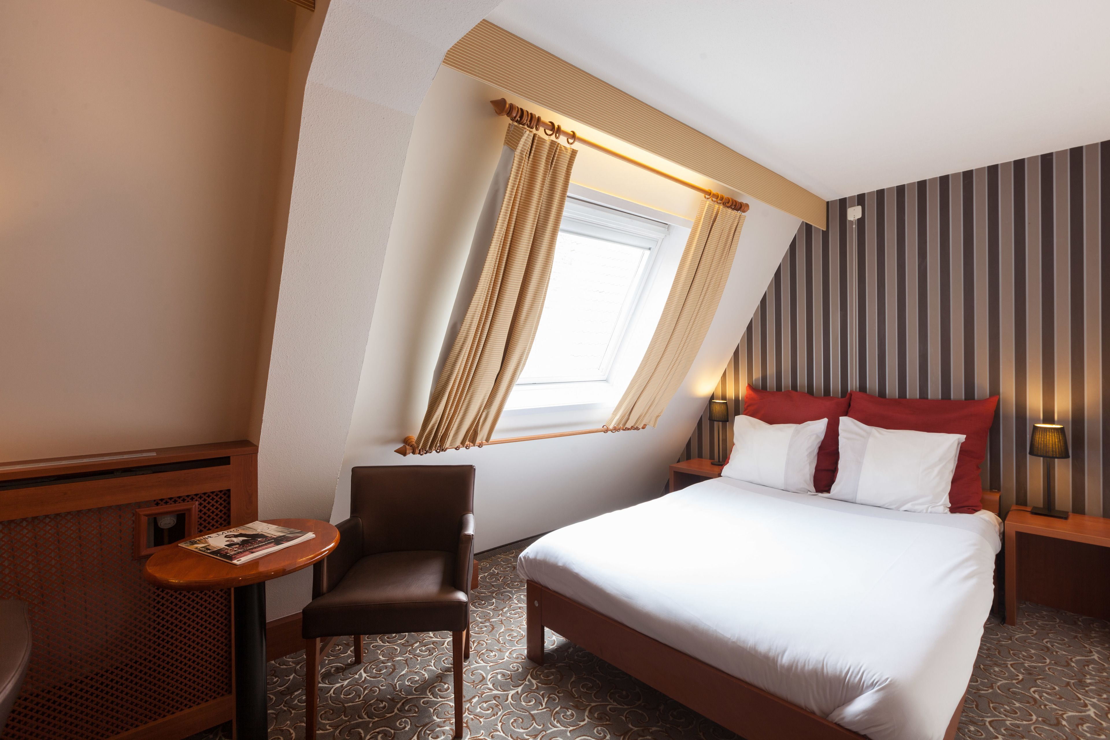 A hotel room with a bed, a chair, a small table, a window with curtains, and a striped wall.