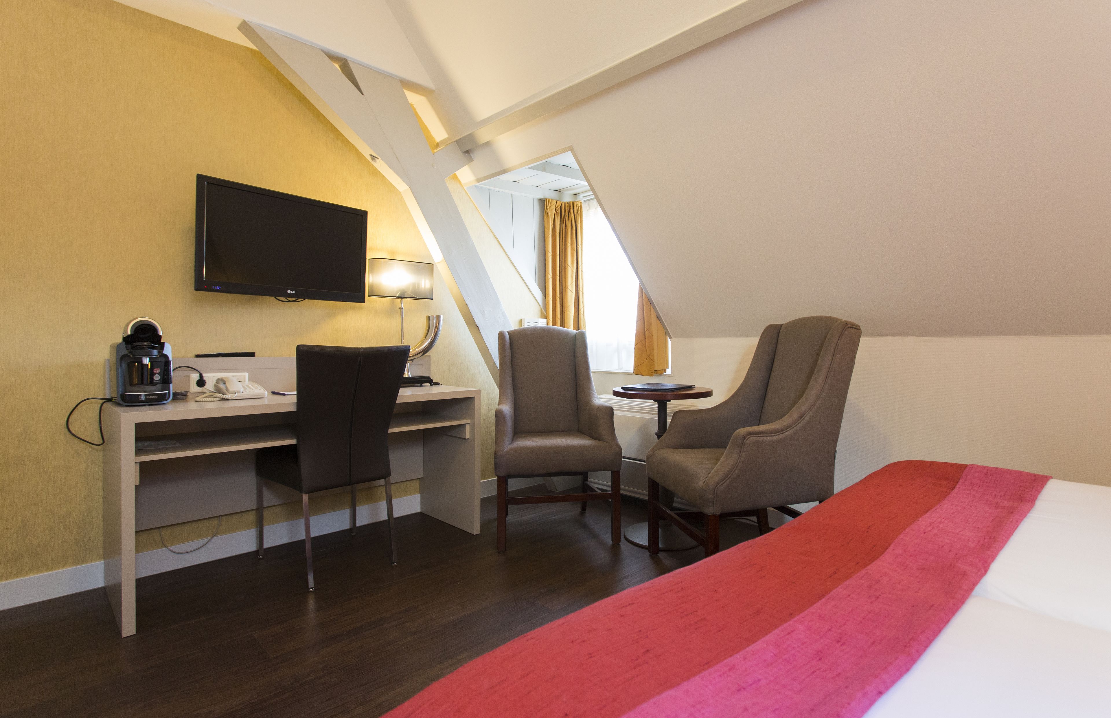 A hotel room with a bed, a desk, two chairs, a television, and a window with curtains.