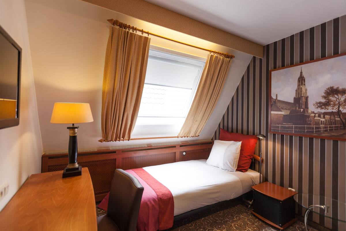A hotel room with a bed, a nightstand, a lamp, a window with curtains, a painting on the wall, and a patterned carpet.