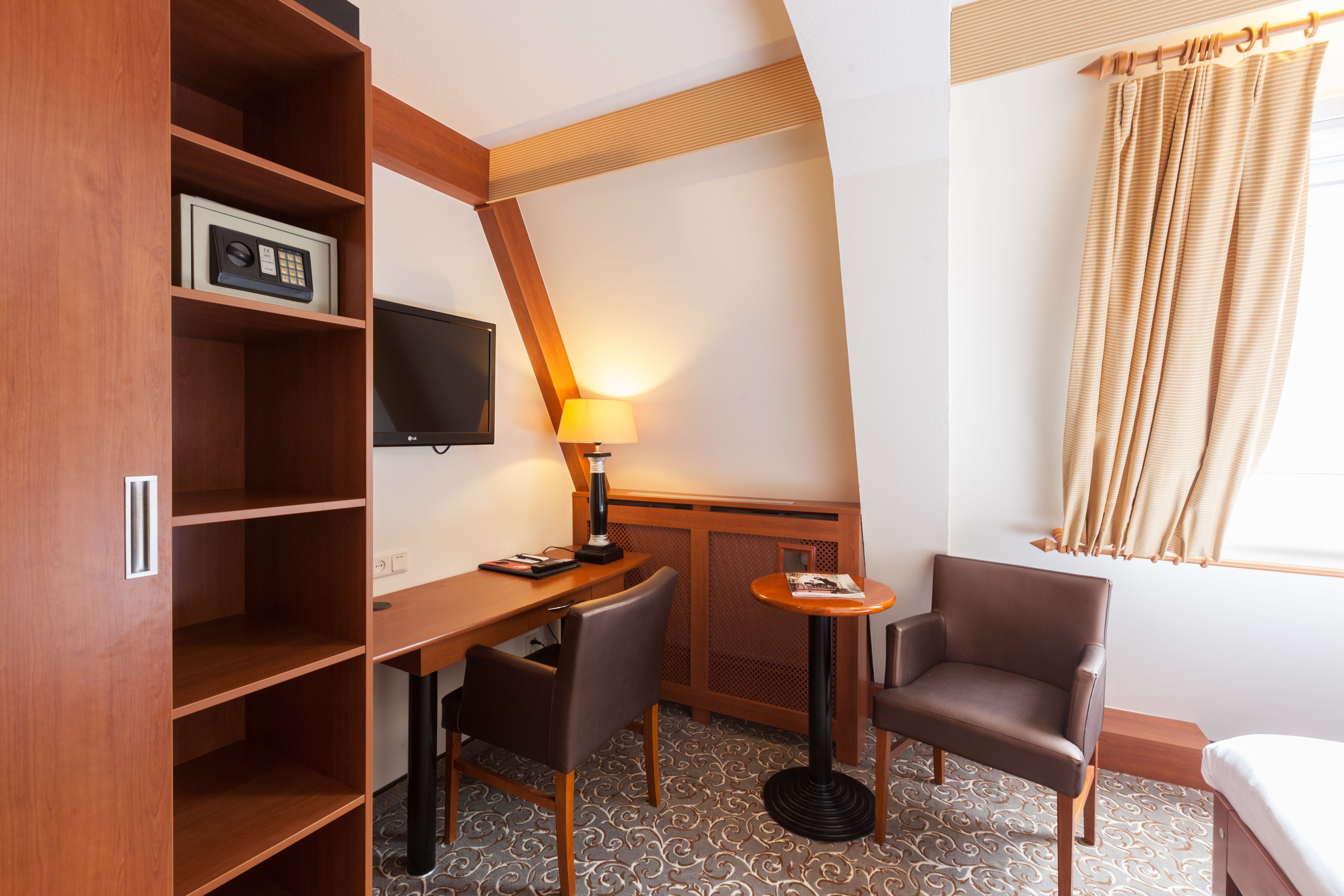A compact hotel room with a wooden desk, two chairs, a television, a lamp, and a window with curtains.