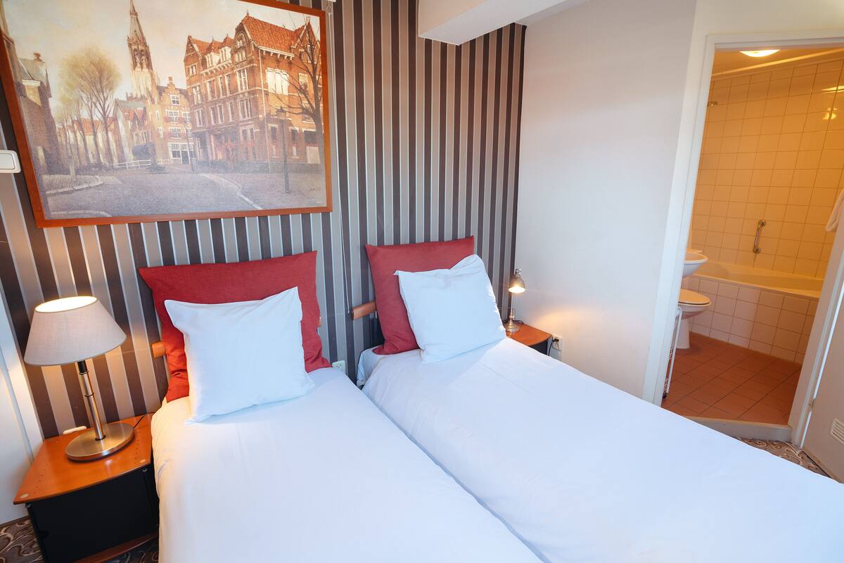 A hotel room with two beds, a nightstand with a lamp, a painting of a street scene, and a view of a bathroom with a bathtub and toilet.