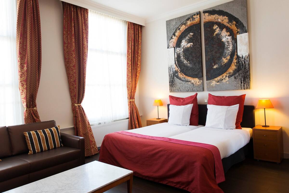 A hotel room with a bed, two bedside tables with lamps, a sofa with striped cushions, and a large abstract painting on the wall.