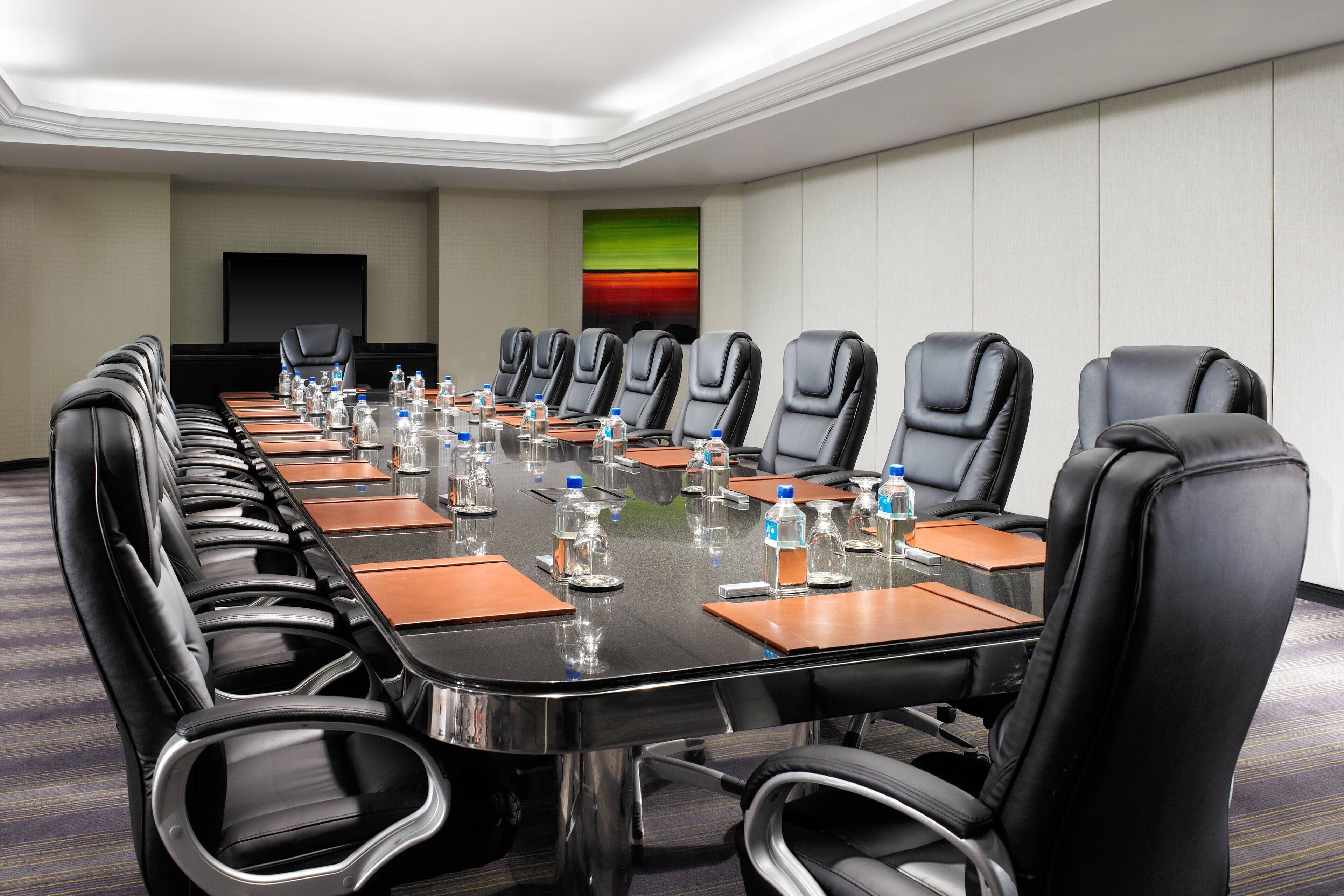 Un salón de conferencias con una mesa larga, sillas de cuero negro, botellas de agua y un televisor de pantalla plana en la pared.