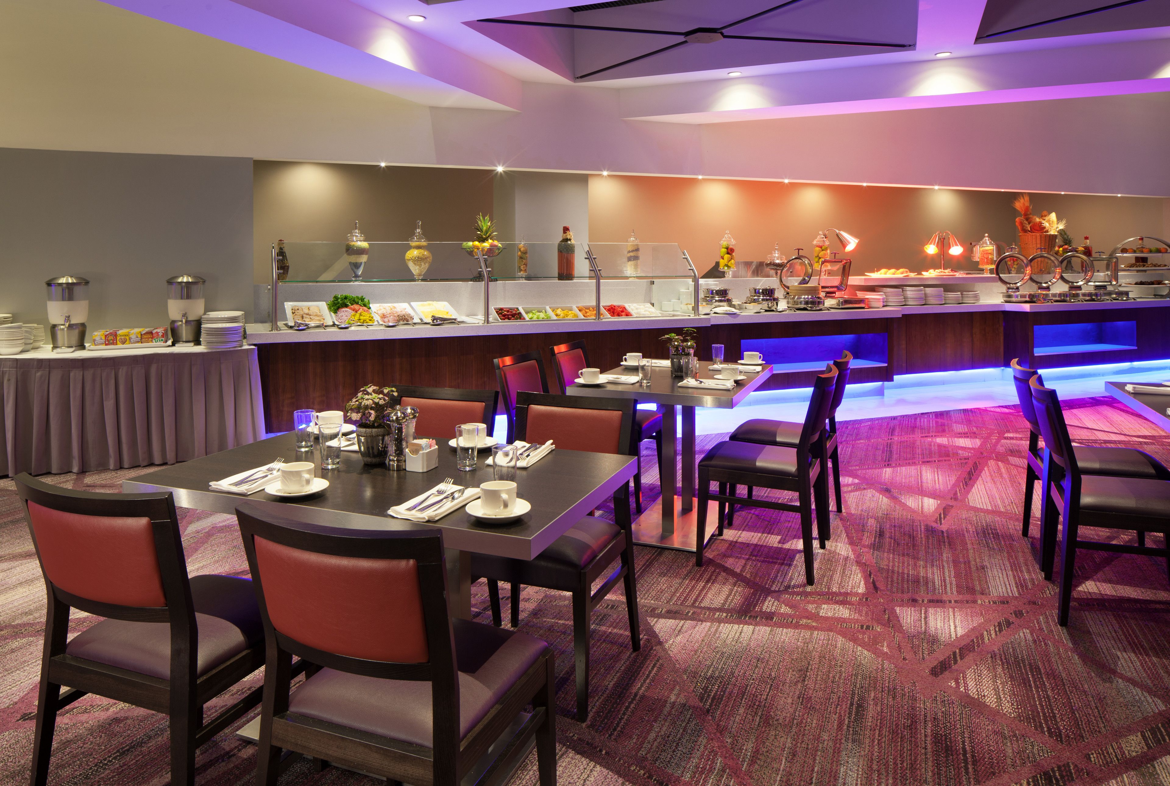 Un comedor de hotel con barra de desayuno buffet, varias mesas y sillas.
