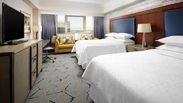 Une chambre d’hôtel avec deux lits, un bureau avec une télévision, un canapé et une grande fenêtre donnant sur la ville.