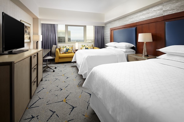 A hotel room with two beds, a desk with a TV, a sofa, and a large window offering a city view.