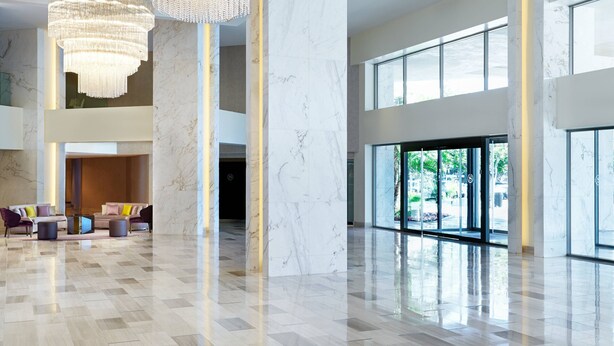 A spacious lobby with a high ceiling, marble columns, and a chandelier. There are seating areas with colorful cushions and a large window offering a view of greenery outside.