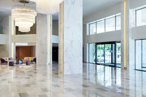 A spacious lobby with a high ceiling, marble columns, and a chandelier. There are seating areas with colorful cushions and a large window offering a view of greenery outside.