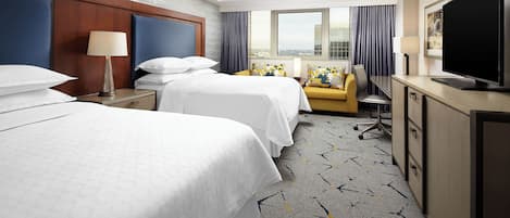 A hotel room with two beds, a desk, a television, and a view of the city through large windows.