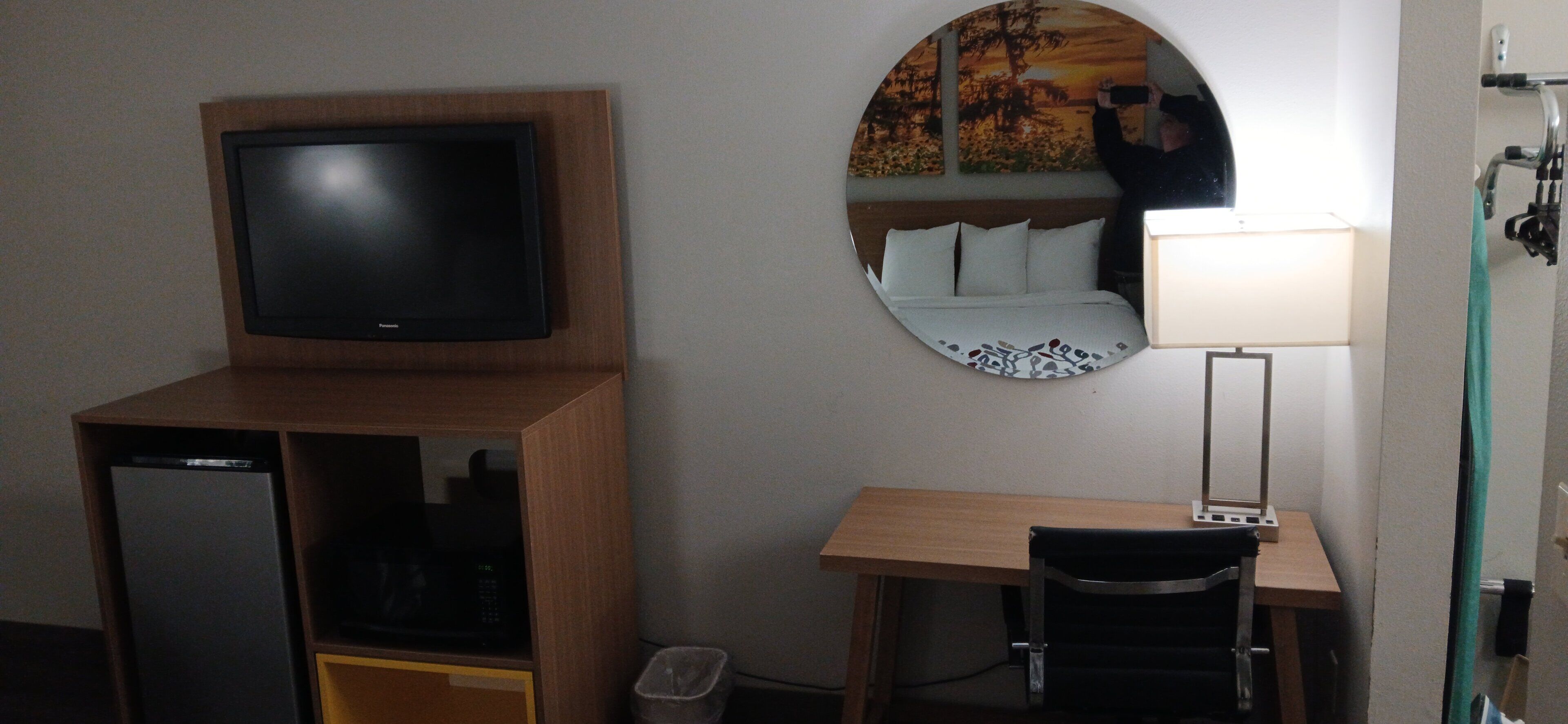 A hotel room with a television on a wooden stand, a desk with a chair, a round mirror, and a bed with white bedding.