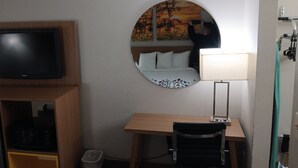 Et hotelværelse med et fjernsyn på et træstel, et skrivebord med stol, et rundt spejl og en seng med hvide sengetæpper.