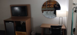 Een hotelkamer met een televisie op een houten standaard, een bureau met stoel, een ronde spiegel en een bed met wit beddengoed.