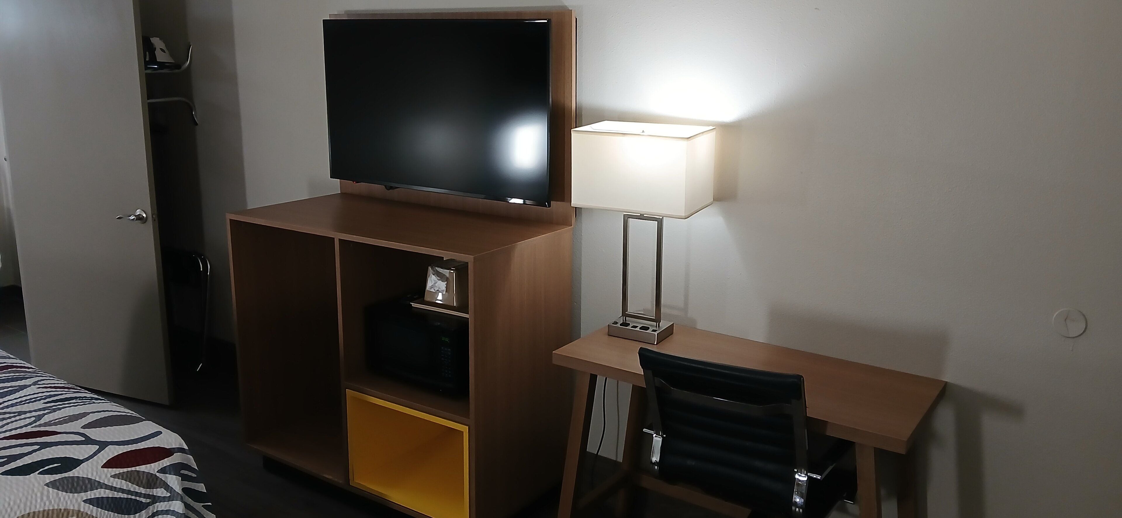 A hotel room with a TV on a wooden stand, a desk with a lamp, a chair, and a bed with a patterned bedspread.