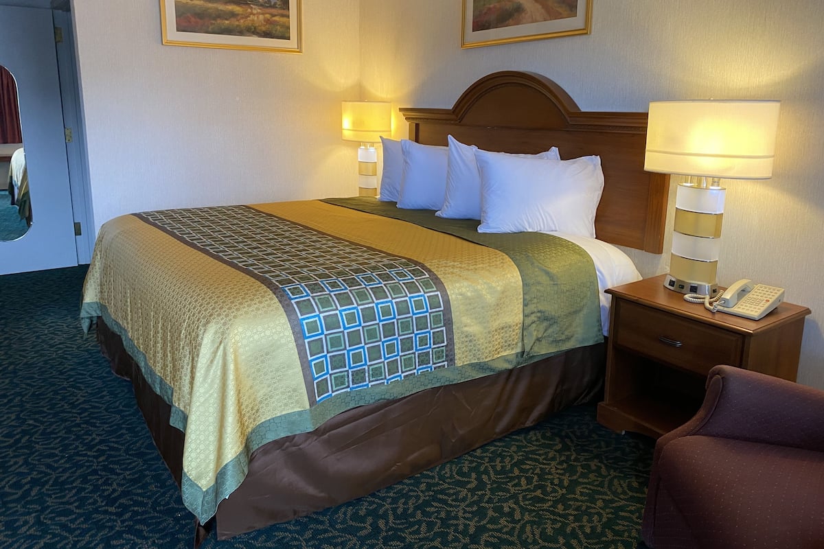 Une chambre d’hôtel avec un grand lit, deux lampes de chevet, un téléphone et deux tableaux représentant des paysages, encadrés et accrochés au mur.
