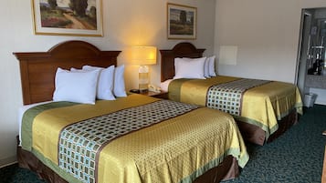 A hotel room with two beds, each with a wooden headboard, white pillows, and a patterned bedspread. There is a nightstand with a lamp and a phone, and a painting on the wall.