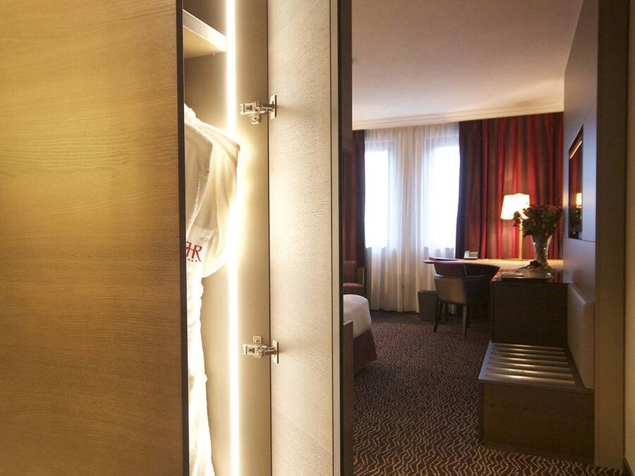 A hotel room with a wooden wardrobe, a desk with a lamp, a chair, a bed, and a patterned carpet.