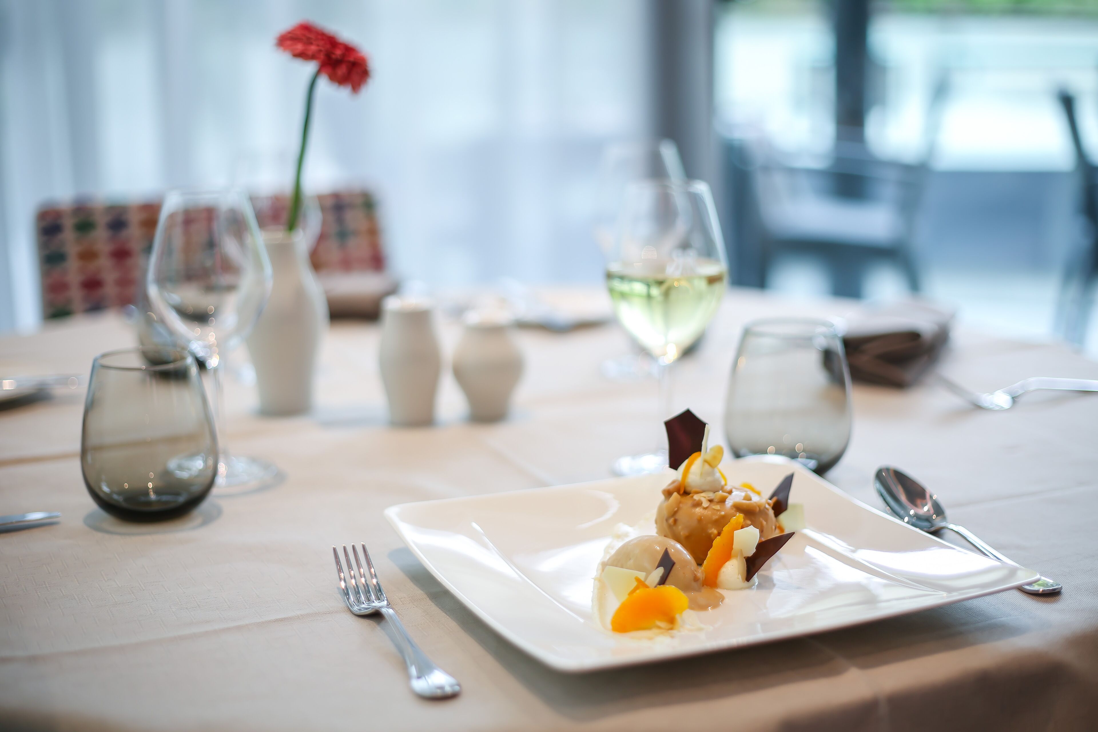 Ein Esstisch mit einem weißen Teller Essen, einer Vase mit einer roten Blume und mehreren Weingläsern.