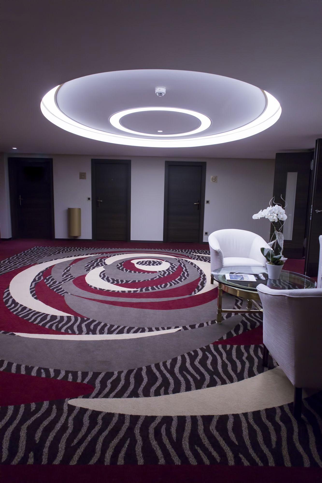 Un hall d’hôtel moderne avec un tapis au motif circulaire, un comptoir d’accueil blanc et un luminaire au plafond.