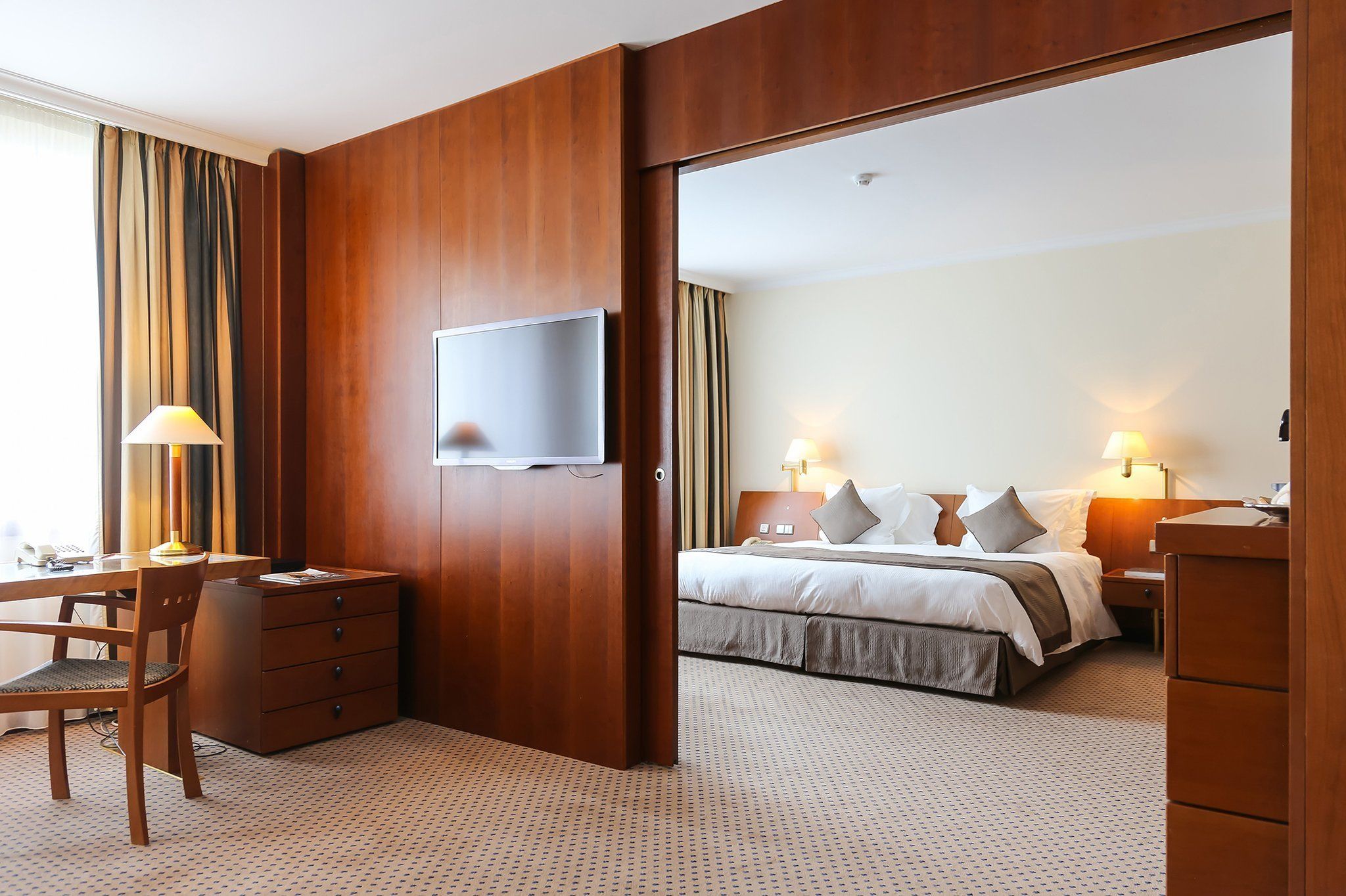 A hotel room with a bed, bedside tables, a desk with a chair, a television, and a wardrobe.