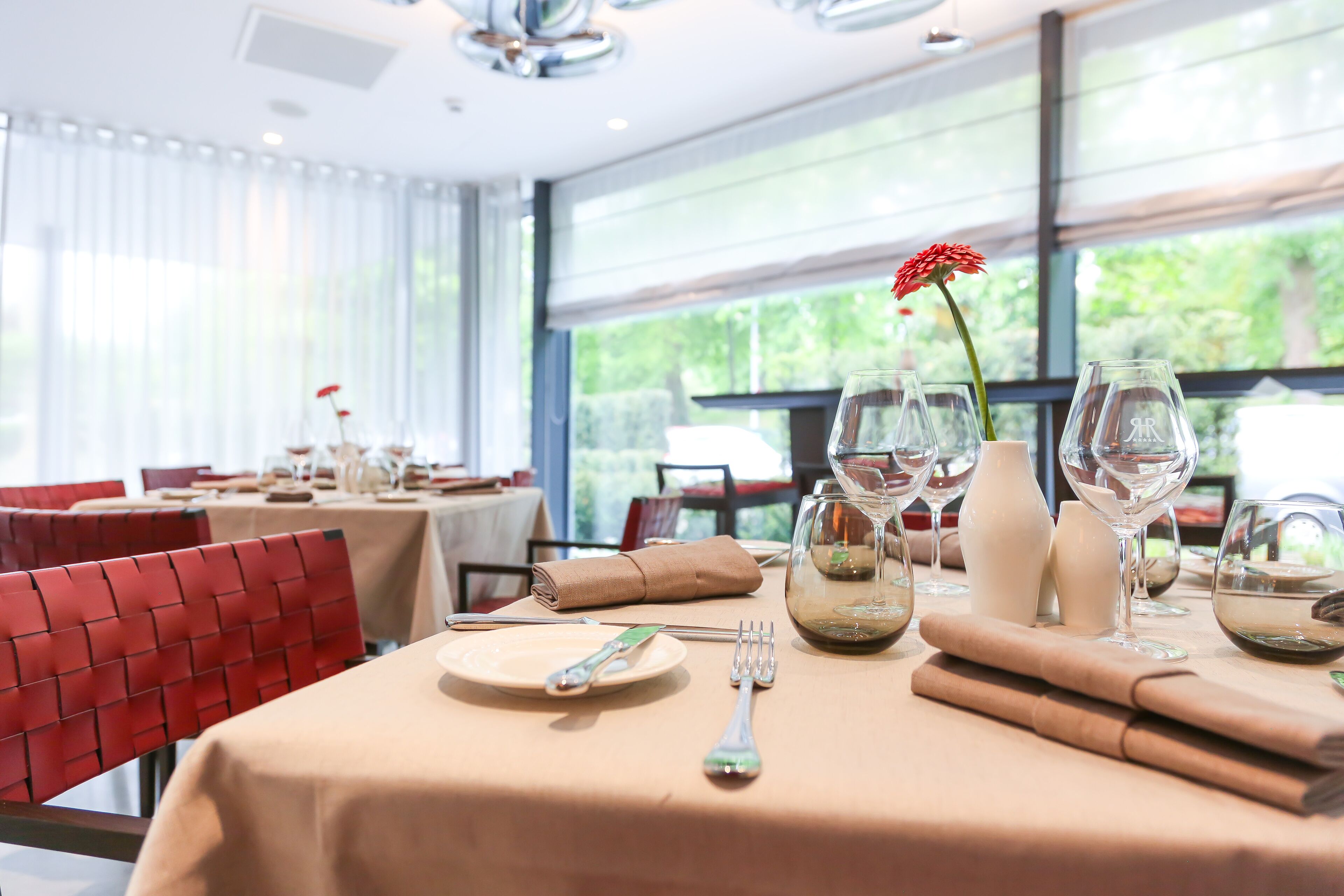 Una mesa de comedor dispuesta con platos, cubiertos y copas de vino, con un centro de mesa de flores rojas.