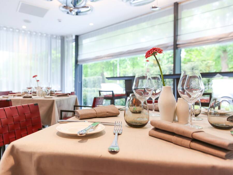 Una mesa de comedor dispuesta con platos, cubiertos y copas de vino, con un centro de mesa de flores rojas.