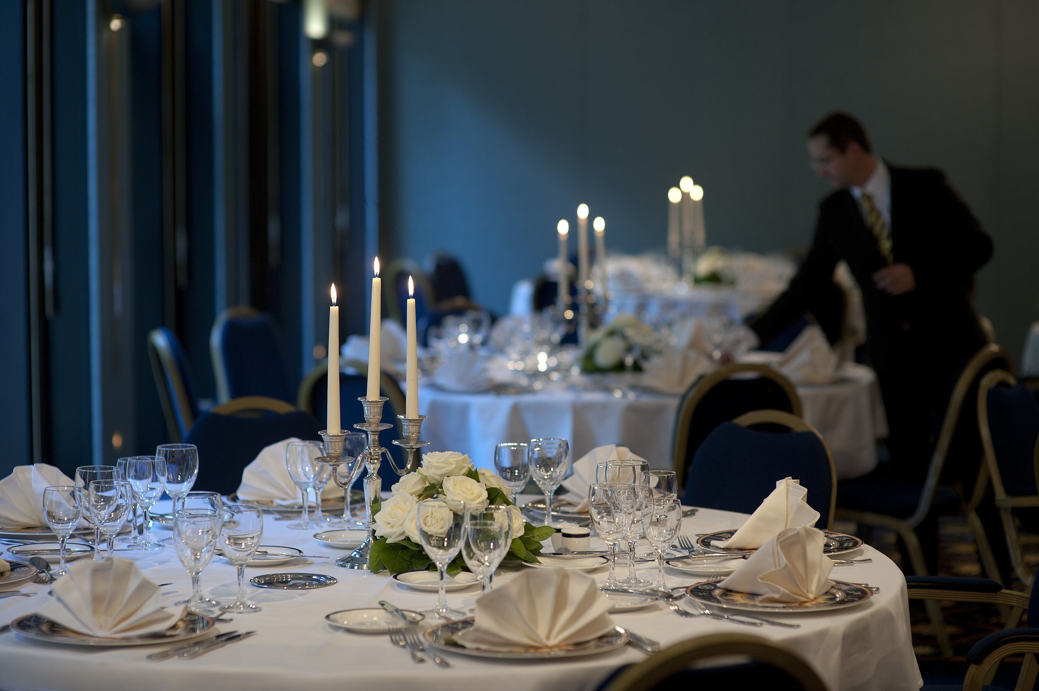 A formal dining room with tables set for a meal, candles, and a waiter in the background.