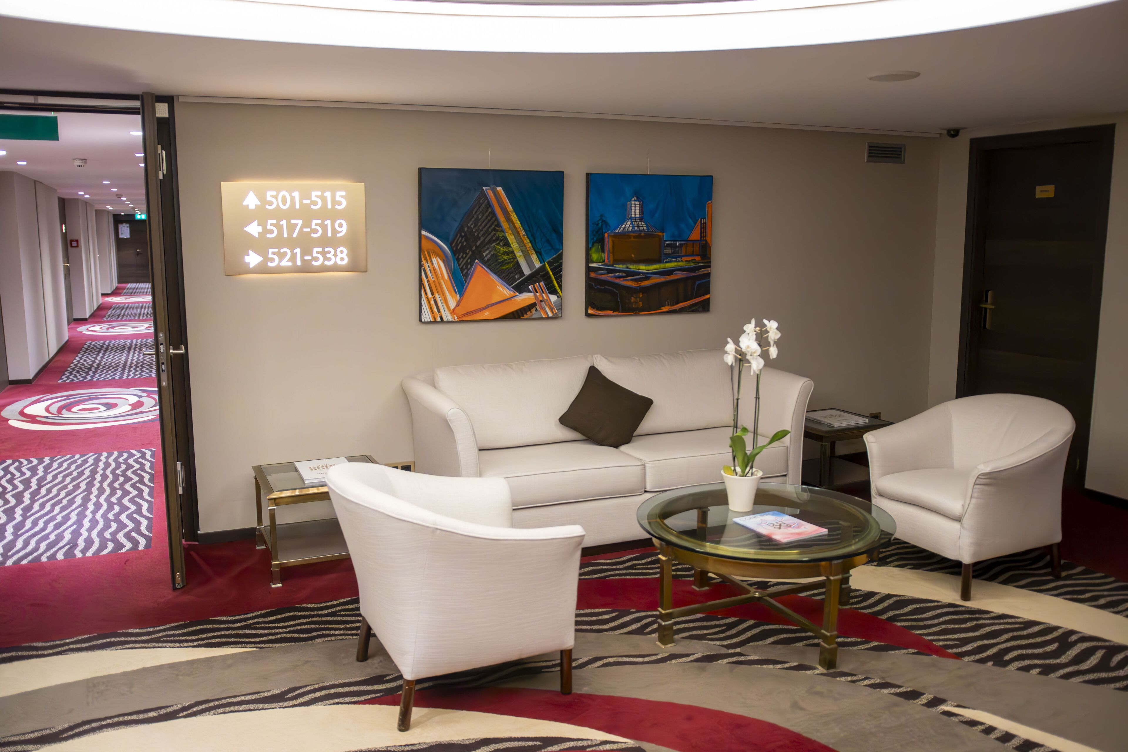 A hotel lobby with a white sofa, two armchairs, a glass coffee table, and two paintings on the wall. There are directional signs above the sofa.
