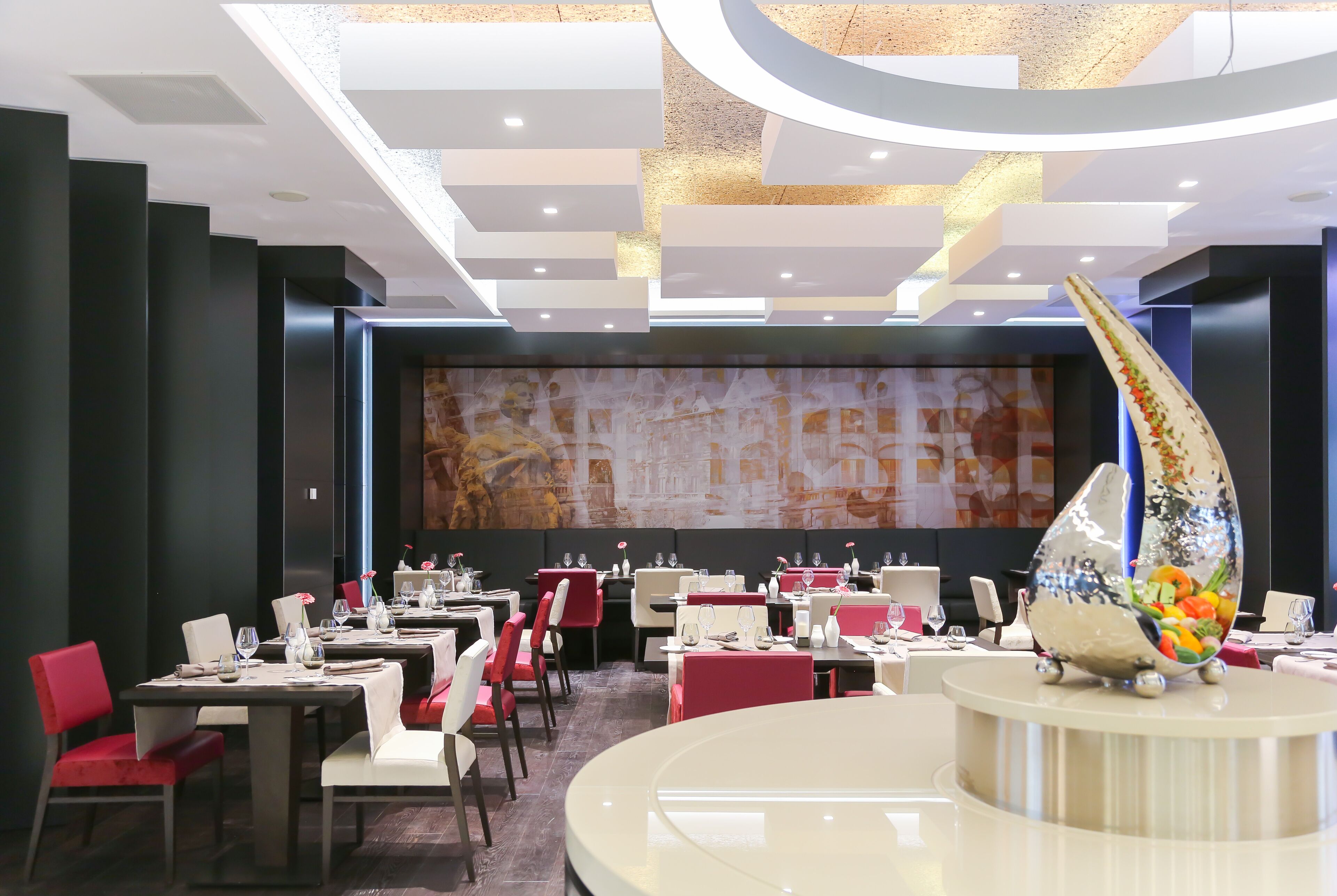 Ein modernes Restaurant mit einem runden Tisch in der Mitte, roten und weißen Stühlen und einem großen abstrakten Wandbild an der Rückwand.
