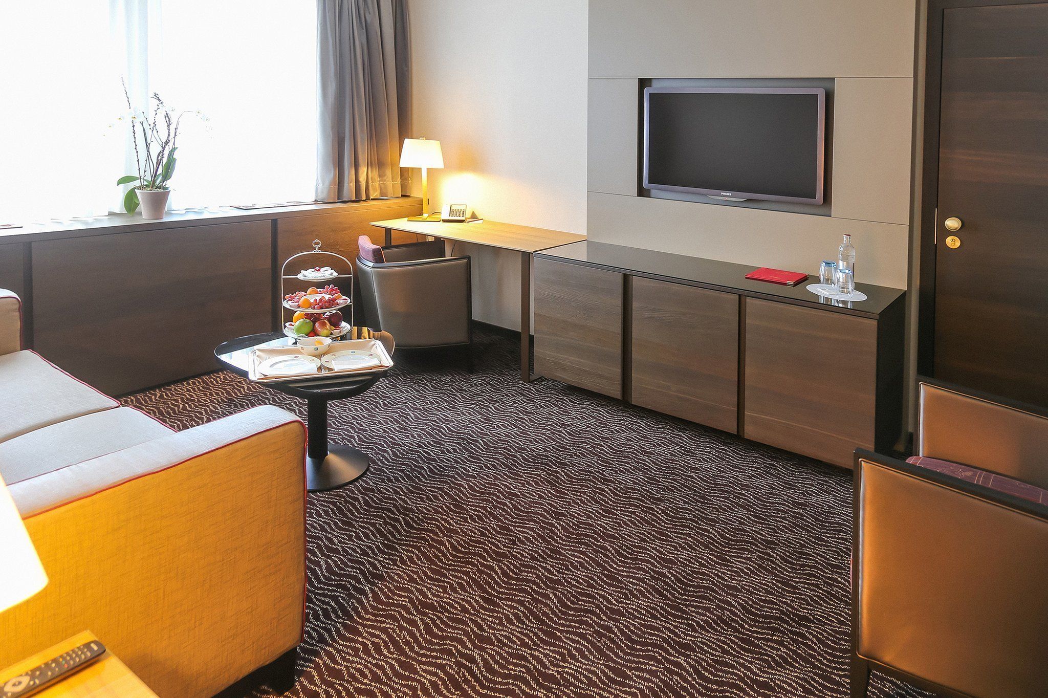 Una camera d'hotel con divano, un tavolino con un vassoio di frutta, una scrivania con lampada, televisione e minibar.