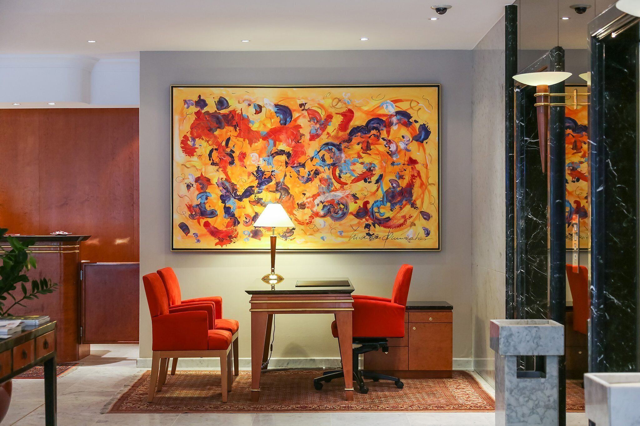 A hotel lobby with a large abstract painting, two red chairs, a desk with a lamp, and a marble column.