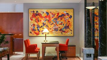 A hotel lobby with a large abstract painting, two red chairs, a desk with a lamp, and a marble column.