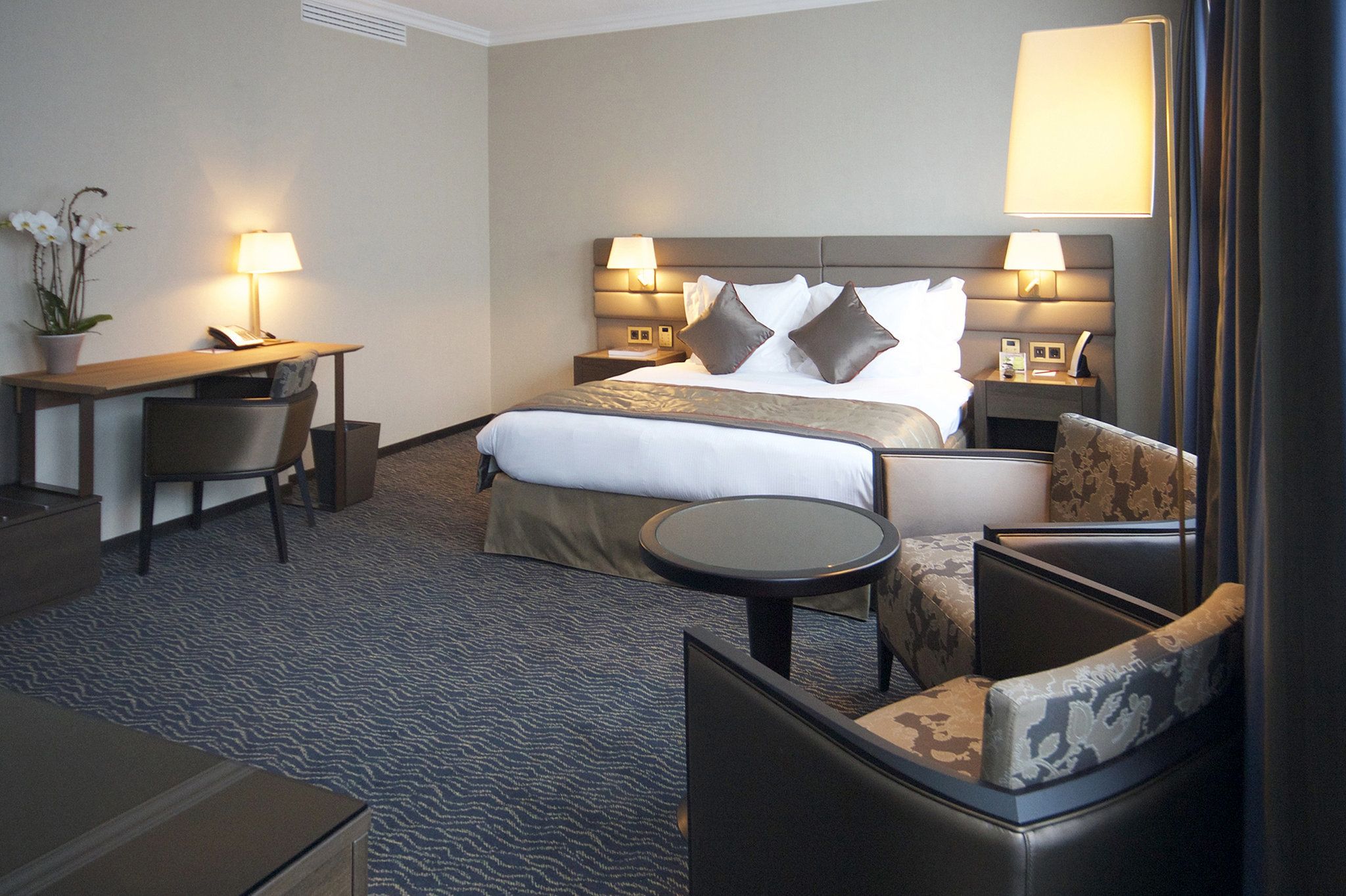 Une chambre d’hôtel avec un grand lit, deux tables de chevet avec des lampes, un bureau avec une chaise et une petite table ronde.