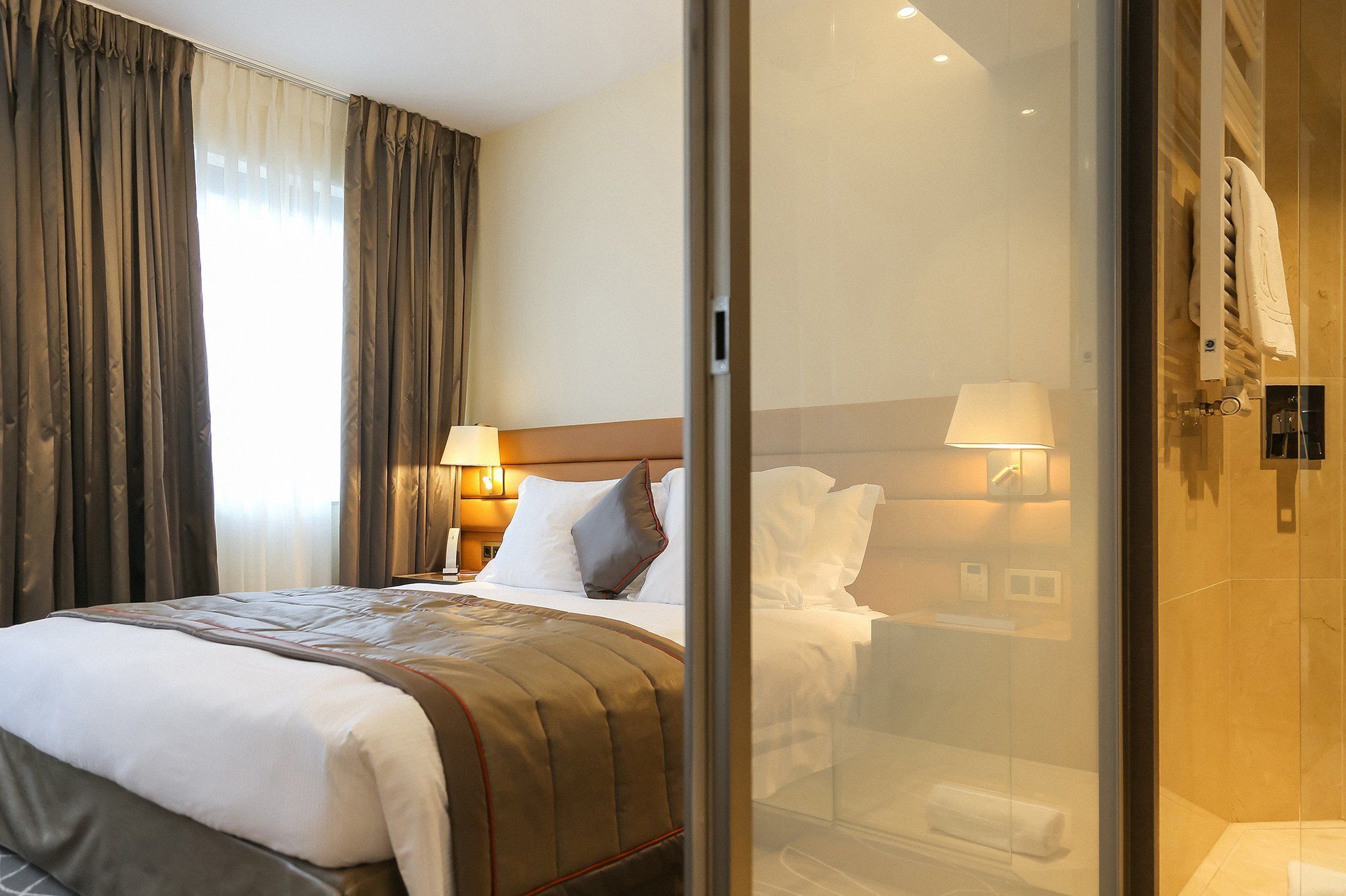 Une chambre d’hôtel avec un lit, une table de chevet avec une lampe, une cabine de douche en verre et une vue sur l’extérieur par la fenêtre.