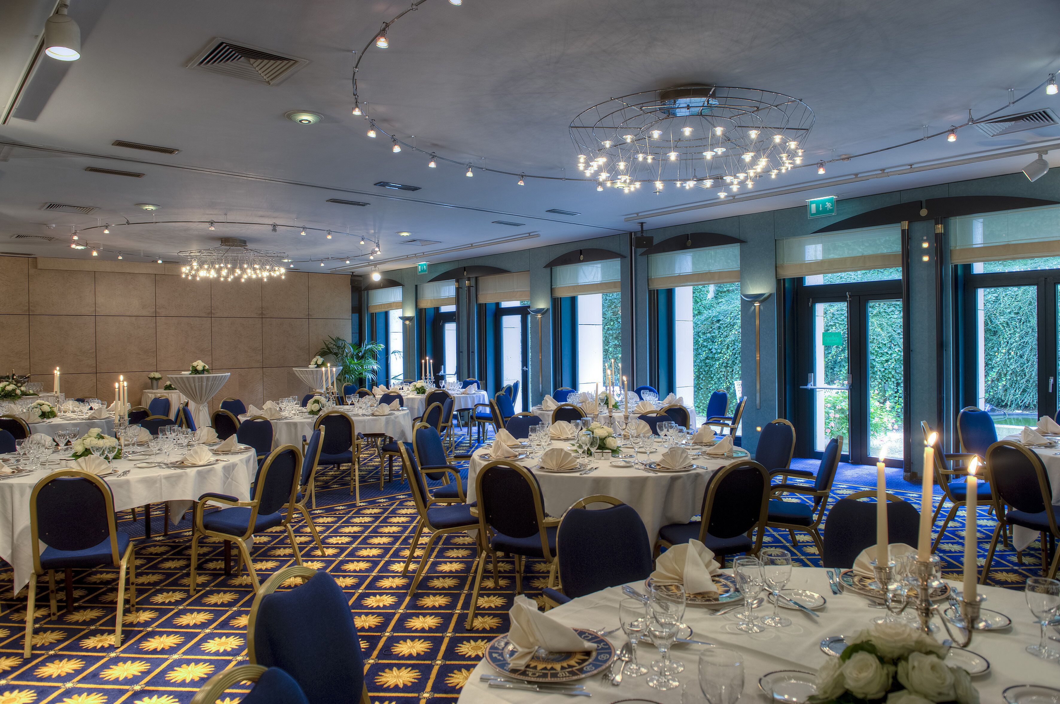 A banquet hall with round tables set for dining, large windows, and a patterned carpet.