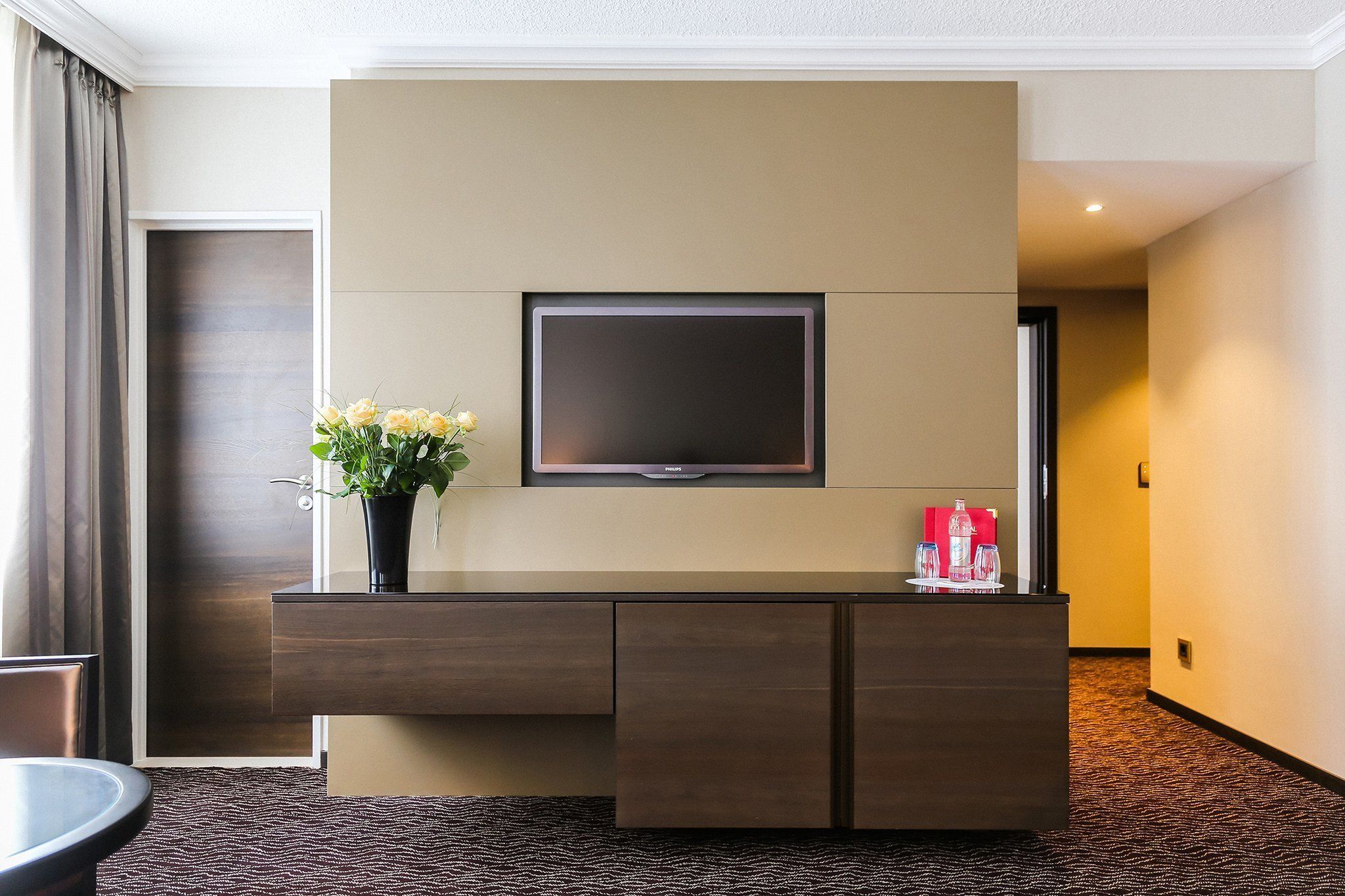 Ein Hotelzimmer mit flachbildfernseher an der Wand, einem dunklen Holzschreibtisch und einem Blumenstrauß sowie einer roten Flasche darauf.