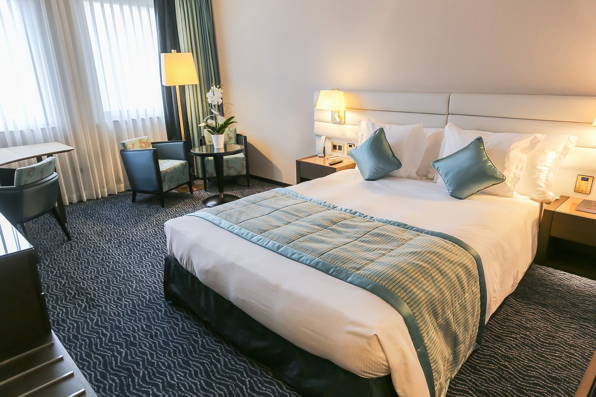 A hotel room with a large bed, a desk, a chair, a small table with a lamp, and a potted plant.